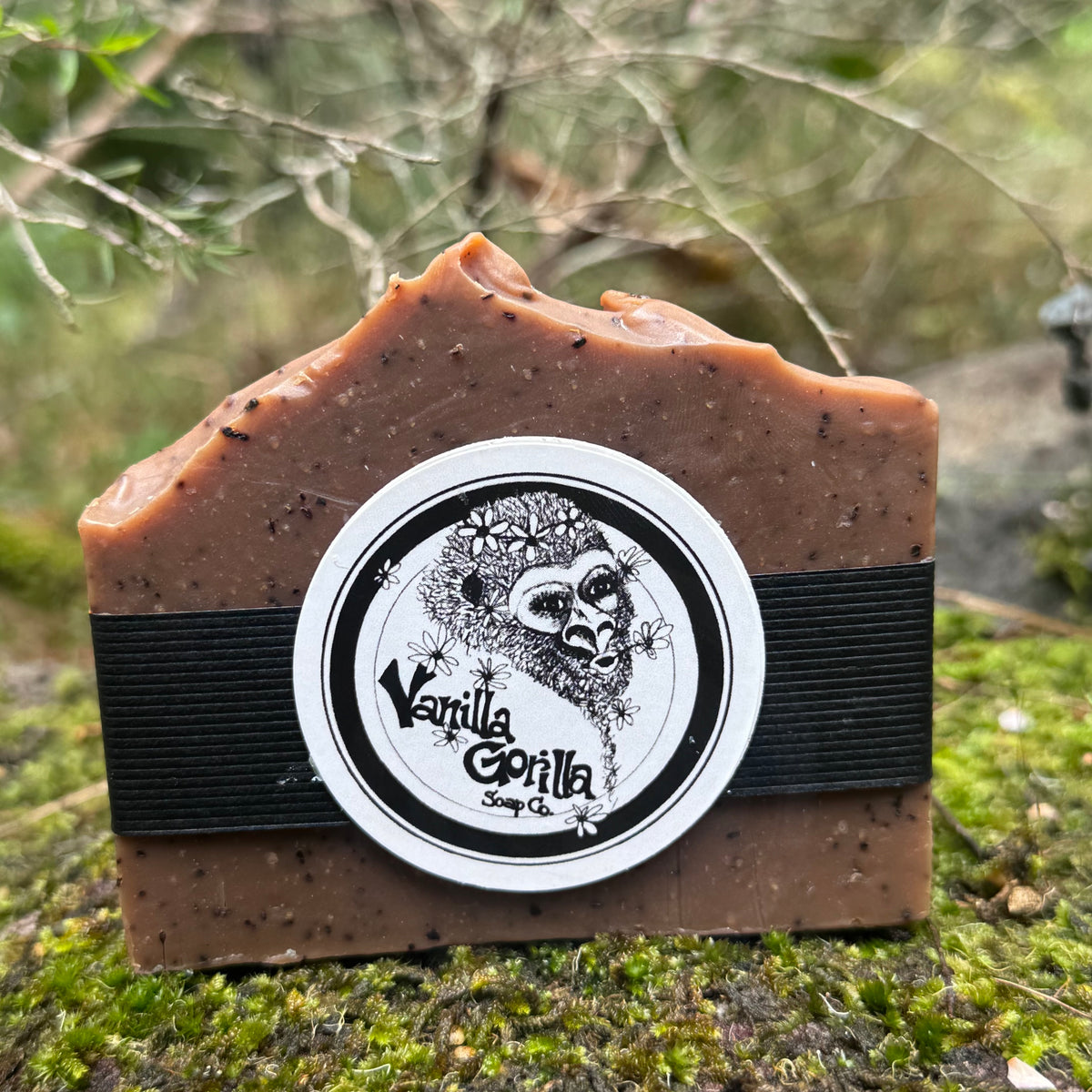 A rectangular bar of brown soap with a jagged top edge, featuring a black elastic band and a round white label that reads 'Vanilla Gorilla Soap Co' with a gorilla illustration. The soap is photographed outdoors on moss with blurred branches and foliage in the background.