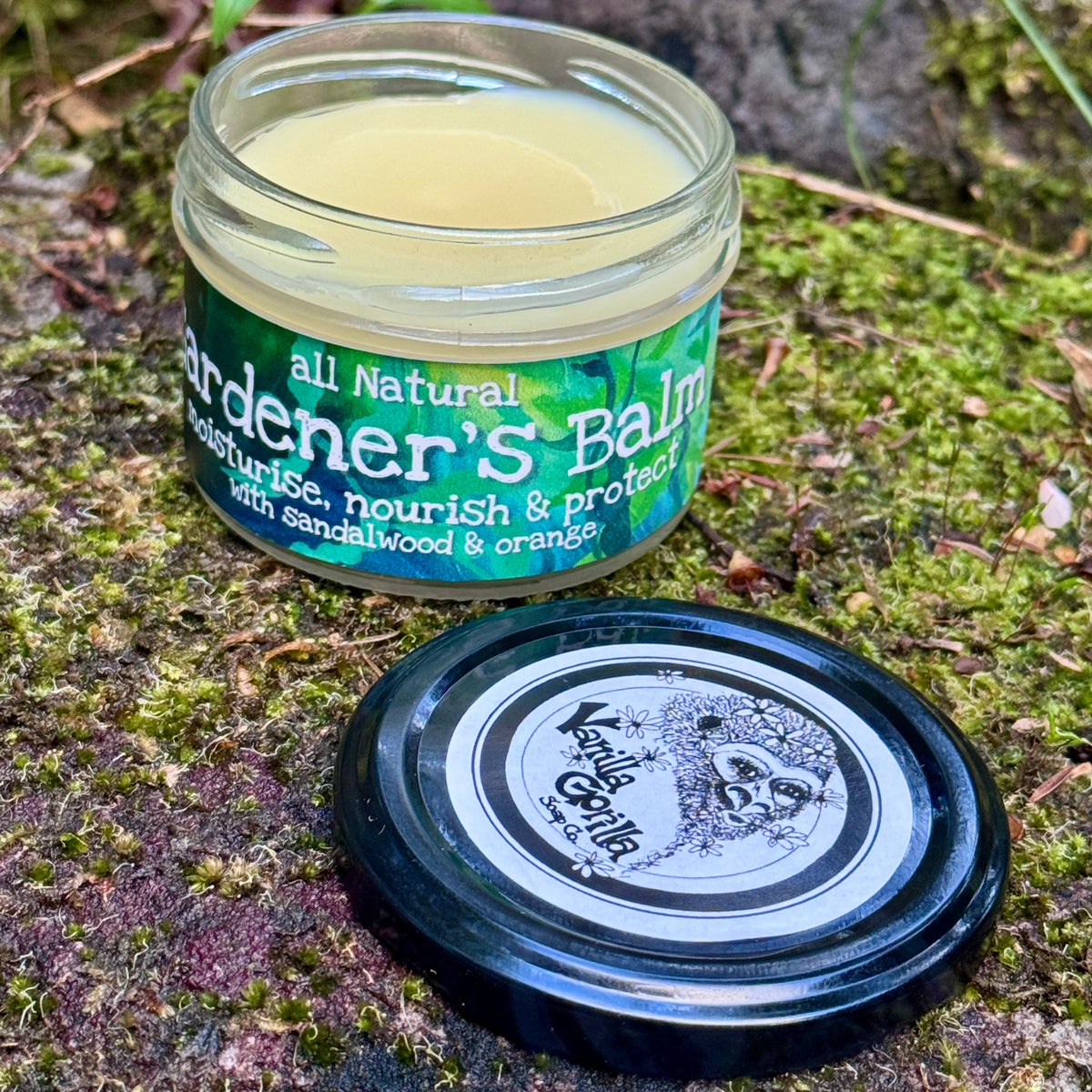 A jar of natural Gardener's Balm with its lid placed in front, set on a mossy outdoor surface. The jar contains a pale yellow balm and has a green label with white text.