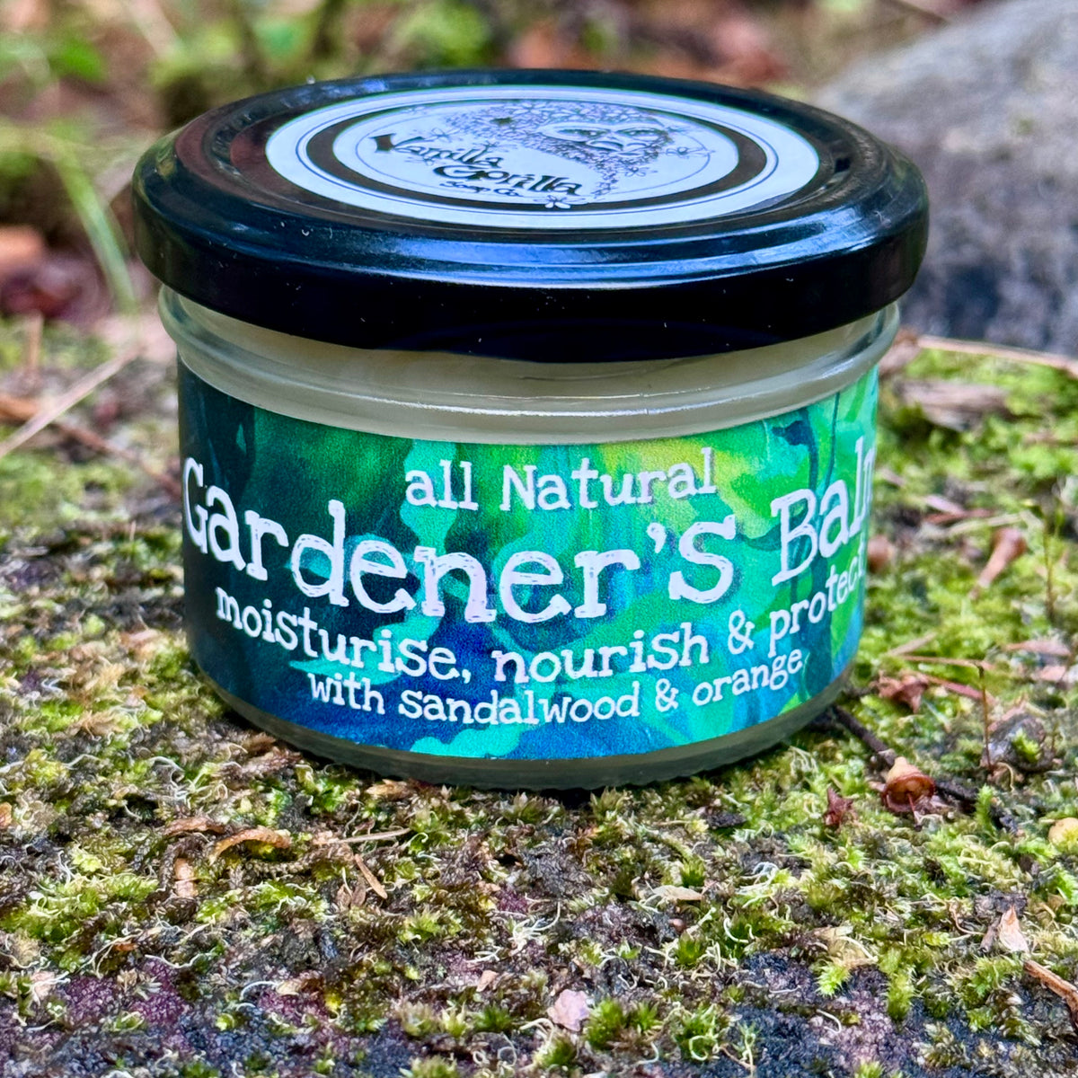 A small jar of Gardener's Balm with a black lid and a green and blue label, placed on mossy ground outdoors. The label reads 'all Natural Gardener's Balm' and 'with sandalwood & orange'.