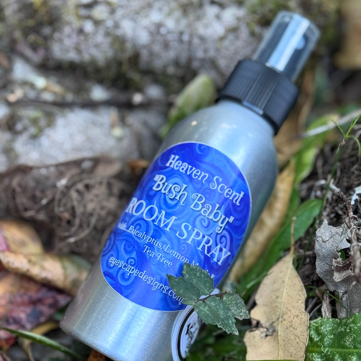 A silver room spray bottle labeled 'Heaven Scent Bush Baby Room Spray' with a blue label, placed outdoors among leaves, moss, and soil.