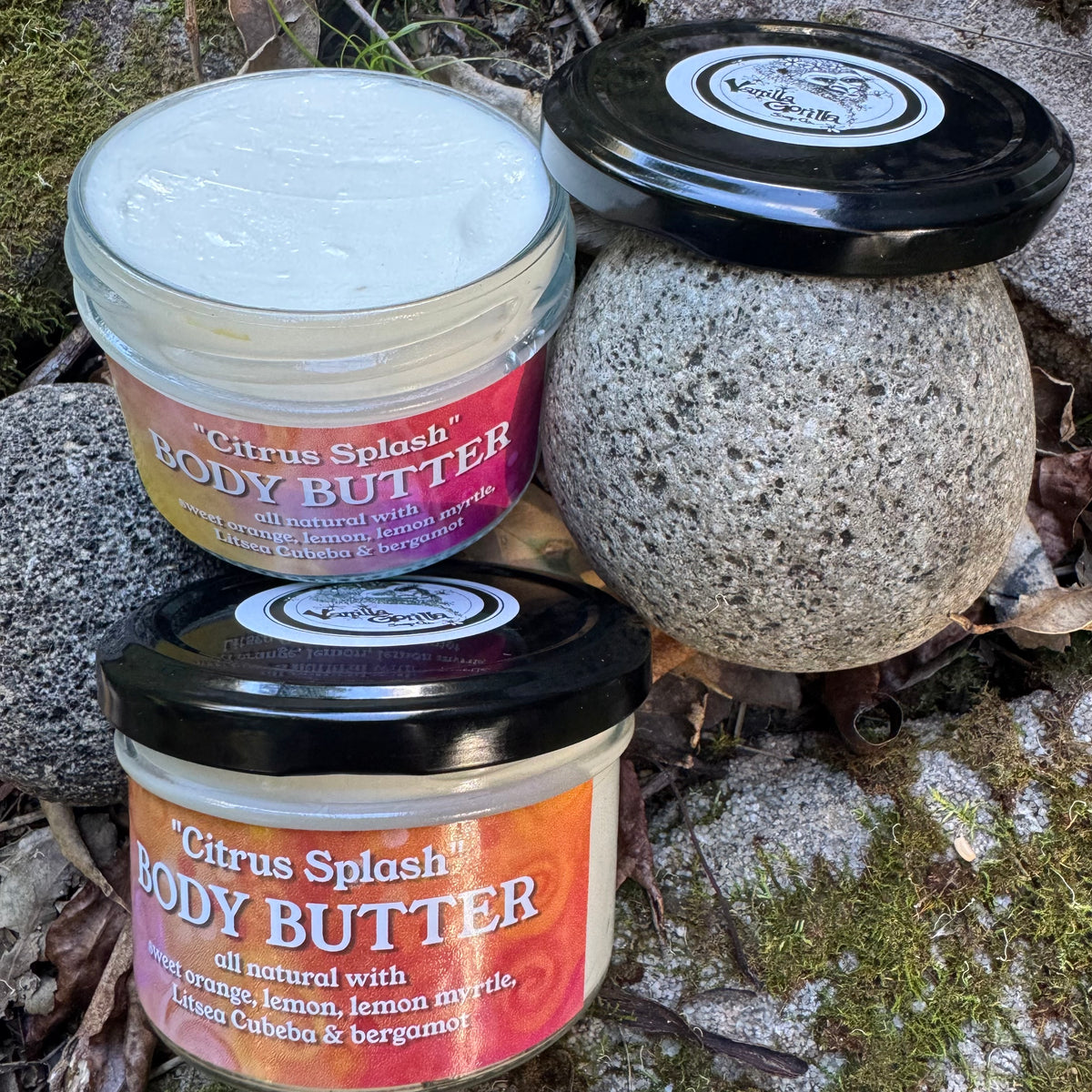 Three jars of Citrus Splash Body Butter arranged outdoors on mossy rocks. One jar is open showing the white cream, one is closed with a black lid and a colorful label, and one is a textured stone jar with a black lid.