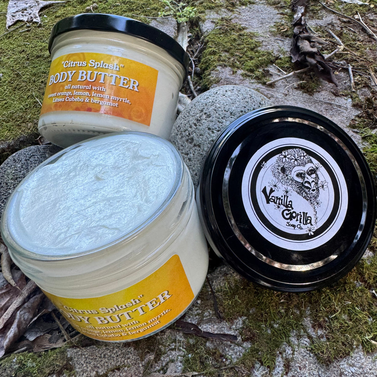 Two jars of body butter and one lid shown on mossy rocks outdoors. The jars are labeled Citrus Splash Body Butter, with one jar open to show the white cream inside and another closed with a black lid featuring a silver and white Vanilla Coaster logo.