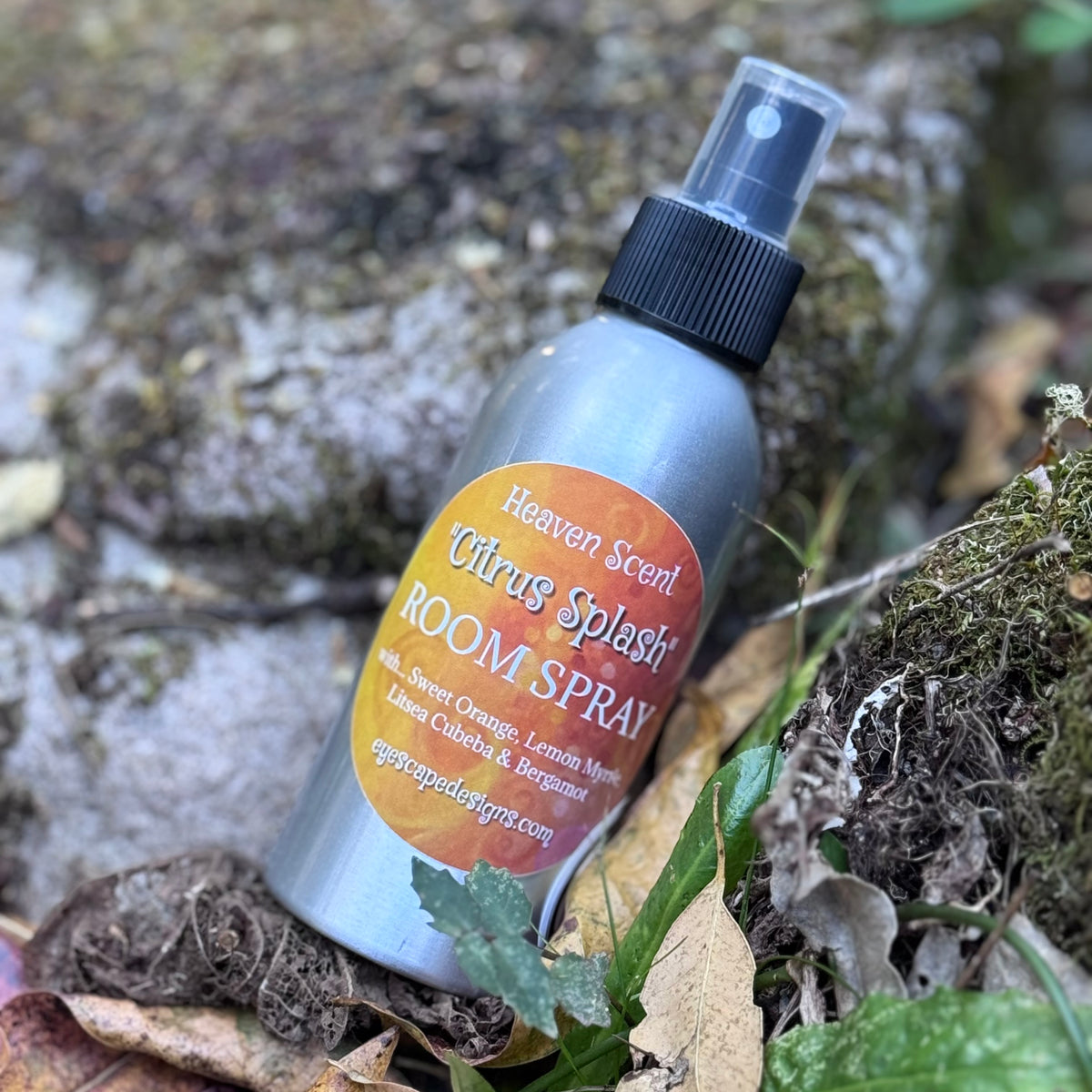 A silver room spray bottle labeled 'Heaven Scent Citrus Scent ROOM SPRAY' resting outdoors among mossy rocks, dry leaves, and green foliage.