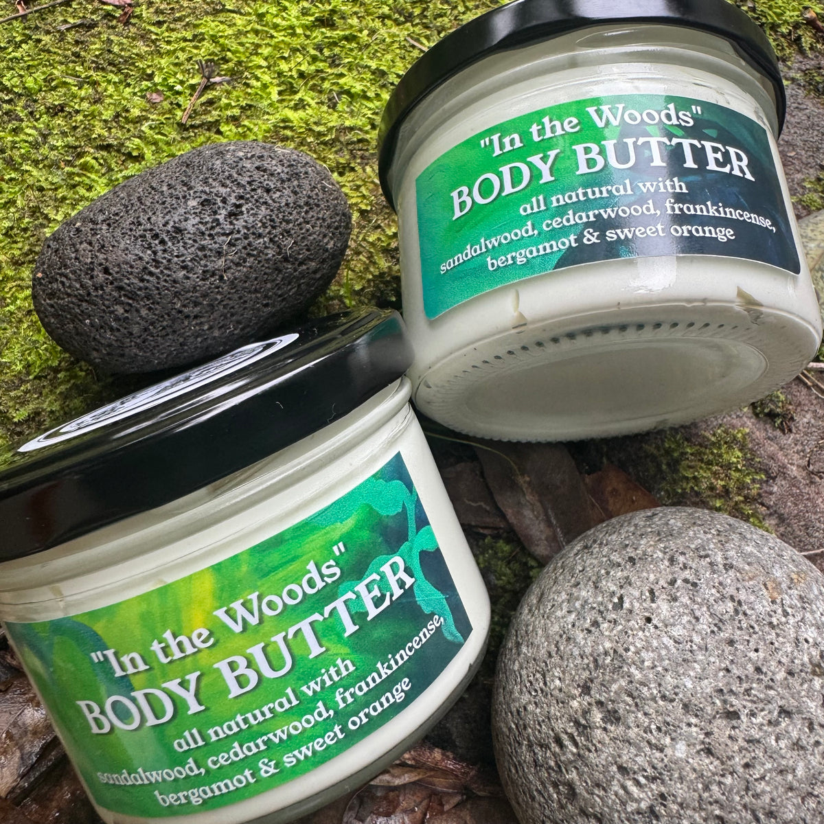 Two containers of 'In the Woods' body butter on a natural background with label sandalwood cedarwood frankincense bergamot sweet orange with stones and moss.