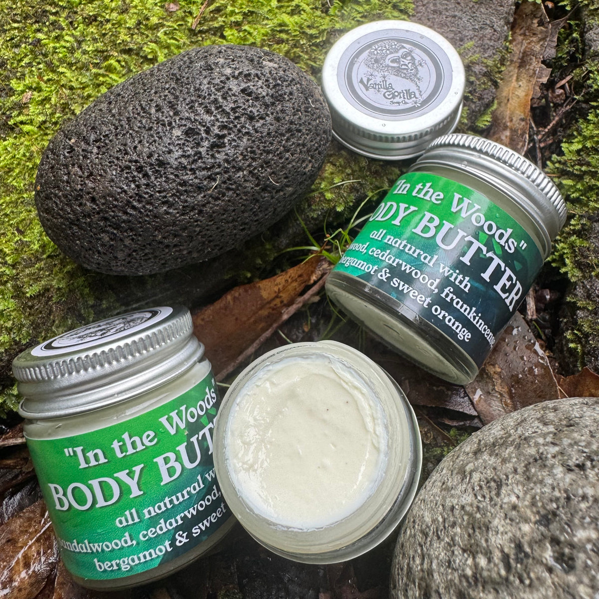 Jars of 'In the Woods' body butter on a natural background with a stone and greenery.