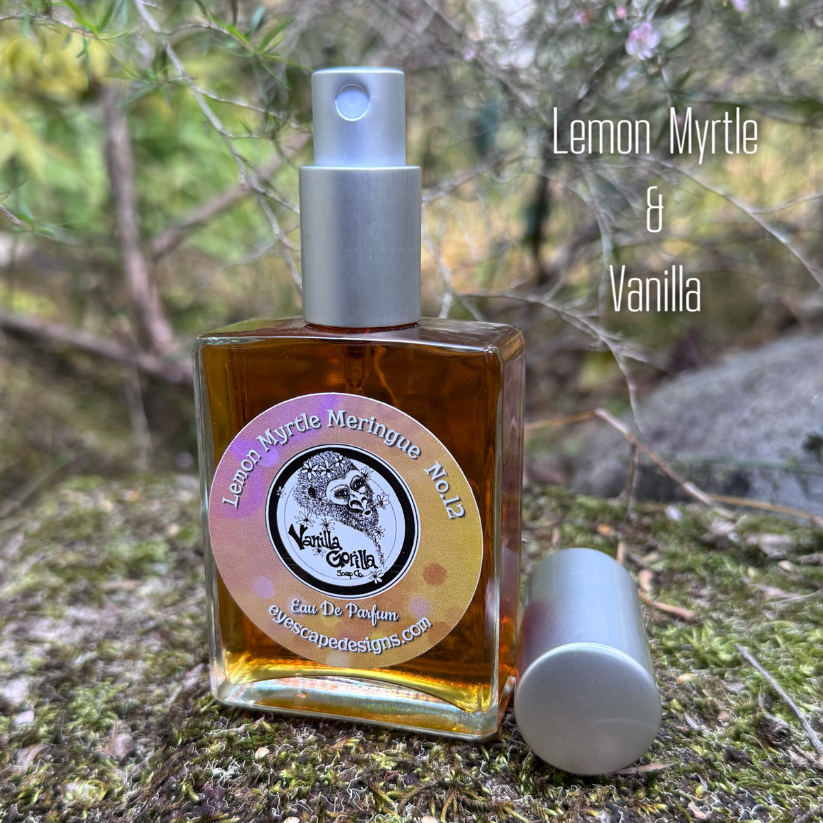A square glass perfume bottle filled with amber liquid sits on mossy ground outdoors, with its silver spray cap removed and placed beside it. The label reads 'Lemon Myrtle & Vanilla' and shows the brand name 'Vanilla Gorilla' with website text.