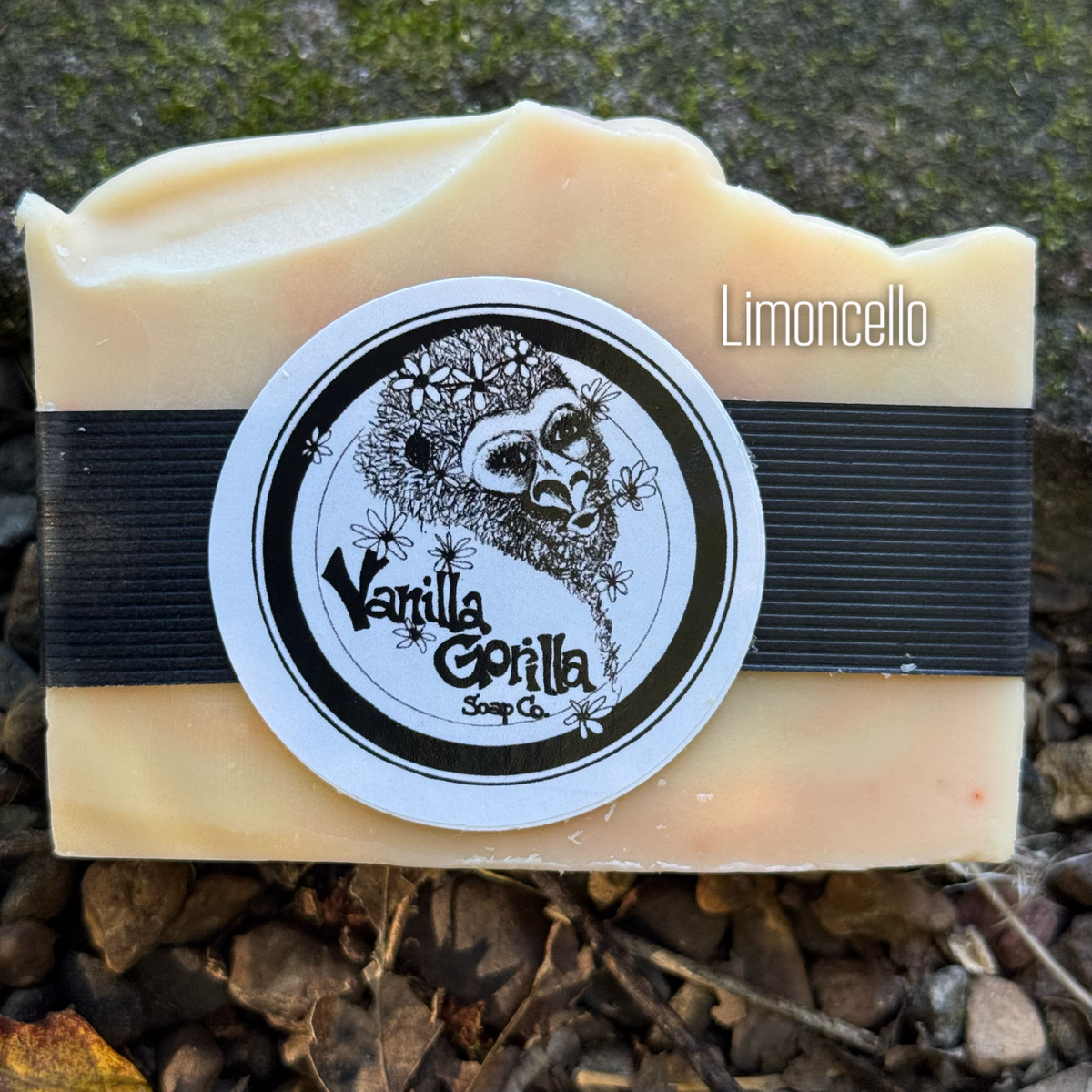 A rectangular bar of soap with a rough, rustic cut, featuring a pale cream-yellow color. It is wrapped with a black elastic band and a round white sticker label that reads 'Vanilla Gorilla Soap Co' with a gorilla illustration.