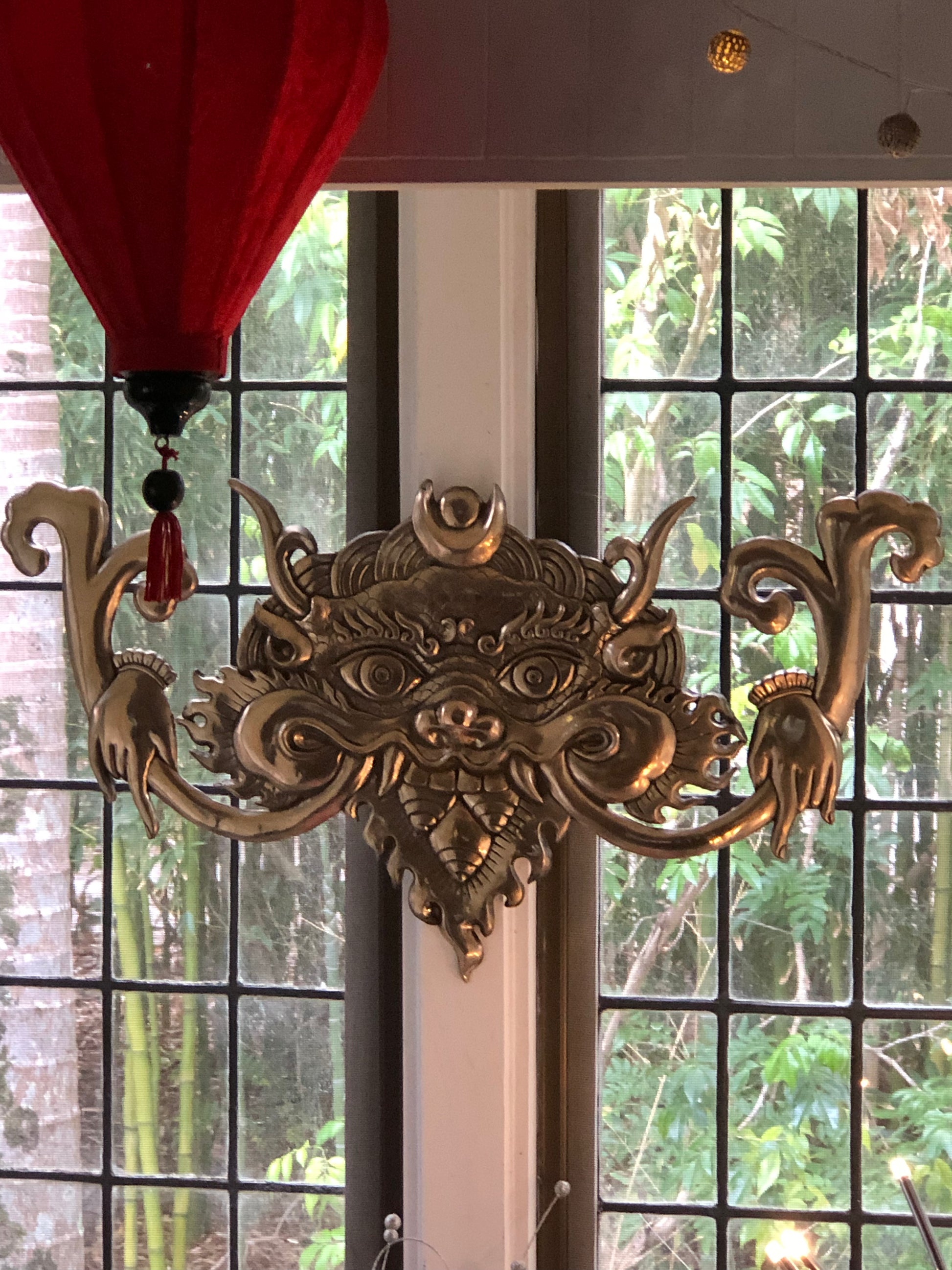 Polished bronze khirtimukha hanging in a bay of windows looking out to a green bush garden