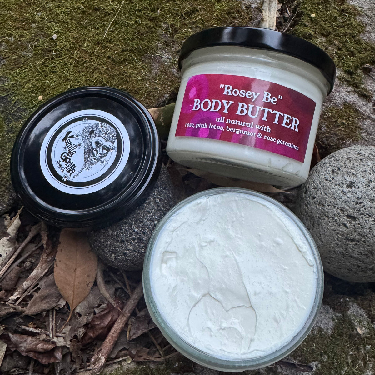A jar of Rosey Be Body Butter shown with its black lid and open container of white body butter on mossy rocks and dried leaves. The jar label reads 'Rosey Be' BODY BUTTER all natural with rose pink lotus, bergamot & rose geranium.