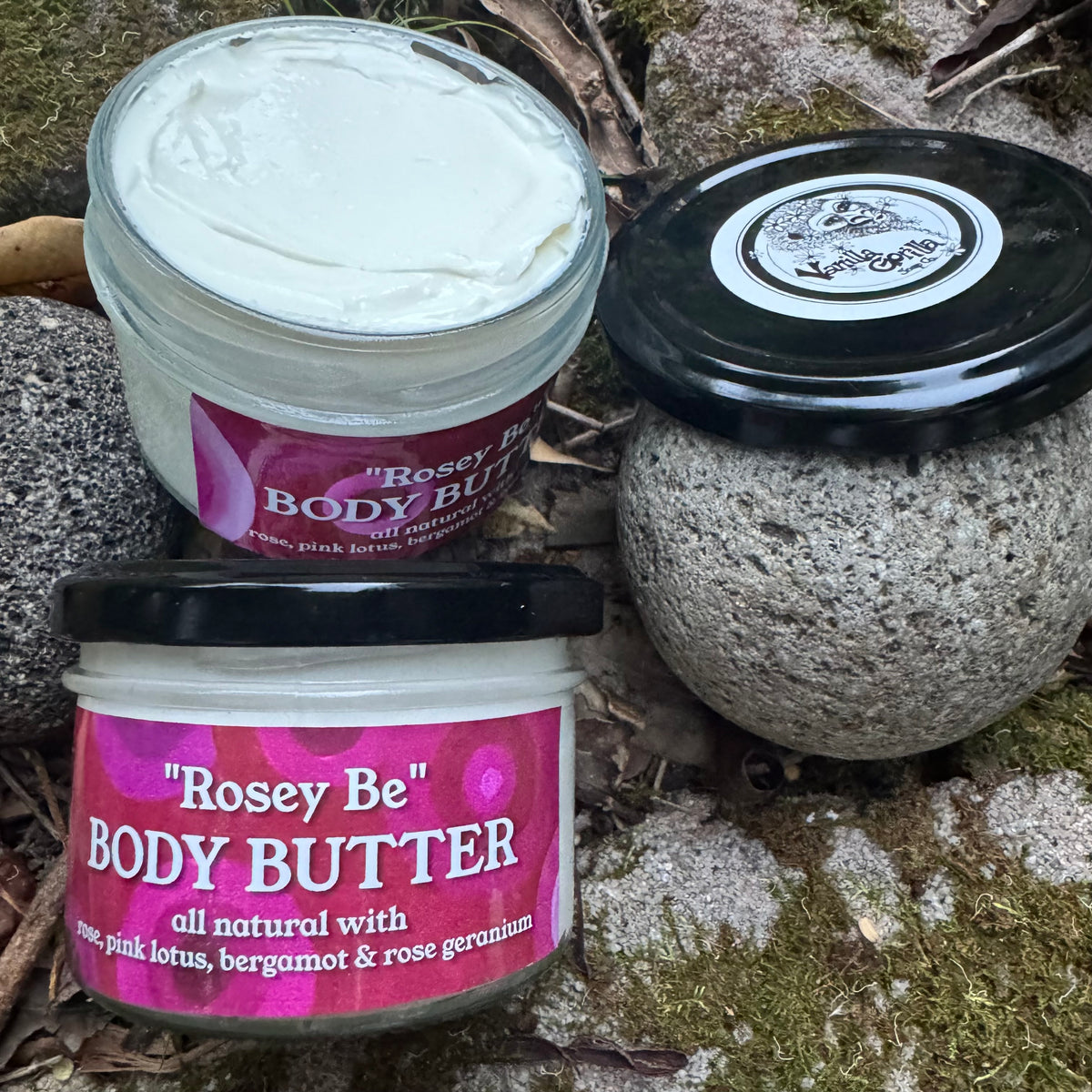 Three jars of Rosey Be Body Butter arranged on mossy rocks outdoors. One jar is open showing the white cream, one is closed with a black lid and a pink label, and one is a textured stone jar with a black lid.
