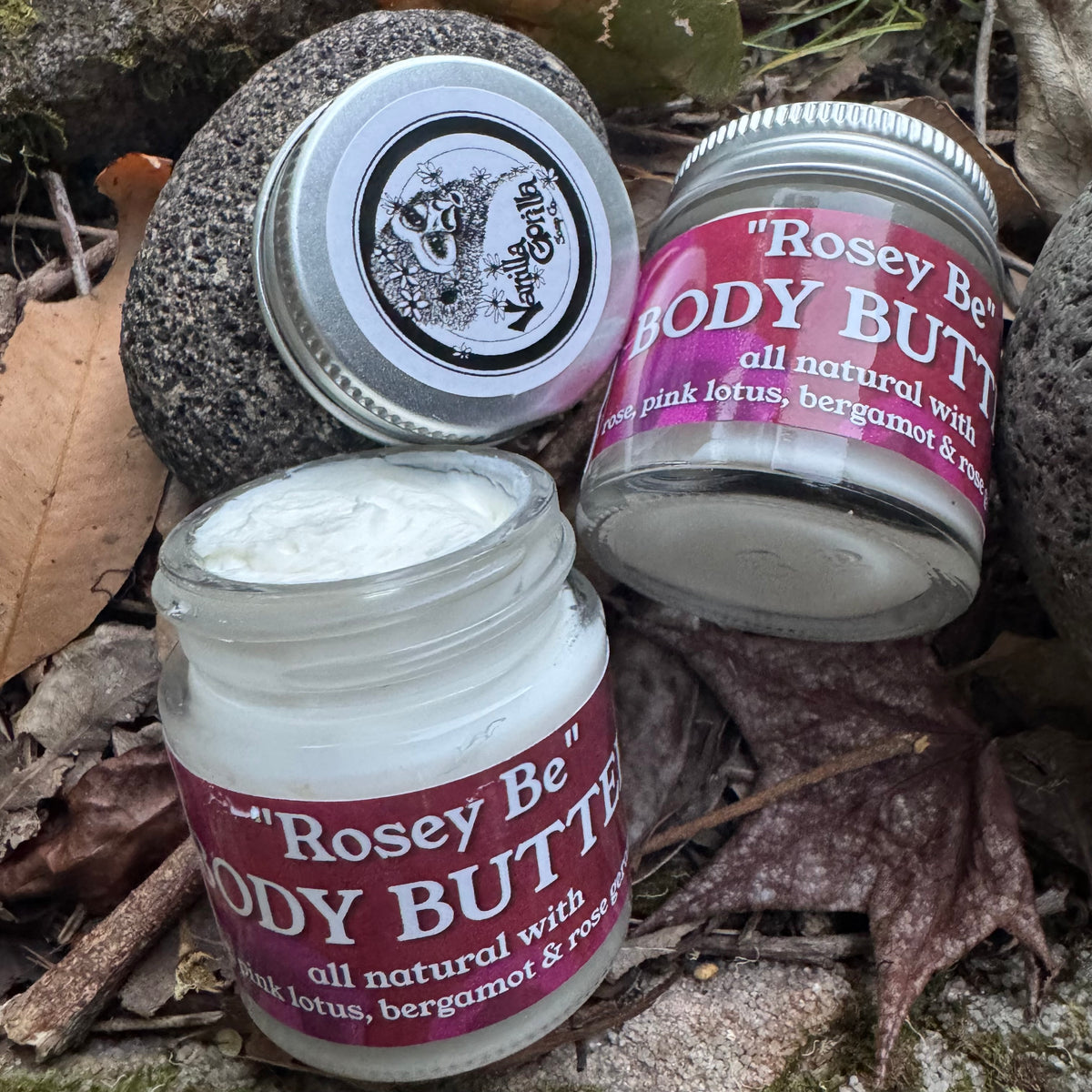 Three jars of Rosey Be Body Butter arranged on a bed of dried leaves and rocks. One jar is open showing white cream, one is closed with its silver lid, and one is upright with a pink label.