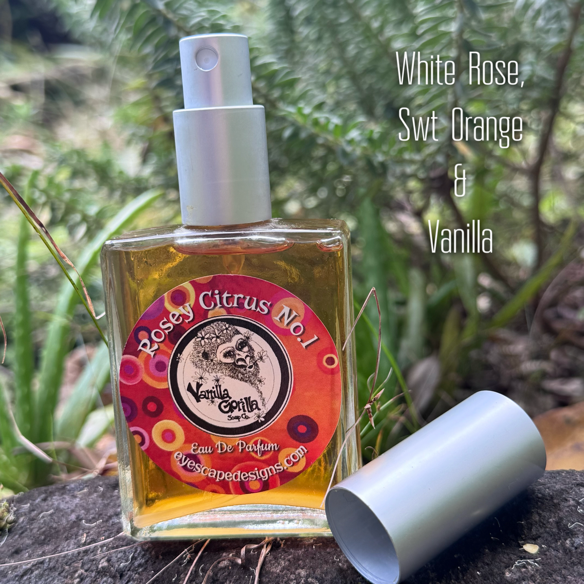 A square glass perfume bottle filled with amber liquid sits outdoors on a dark log with green foliage in the background. The bottle has a silver spray top and a colorful circular label reading 'Rose Citrus No.