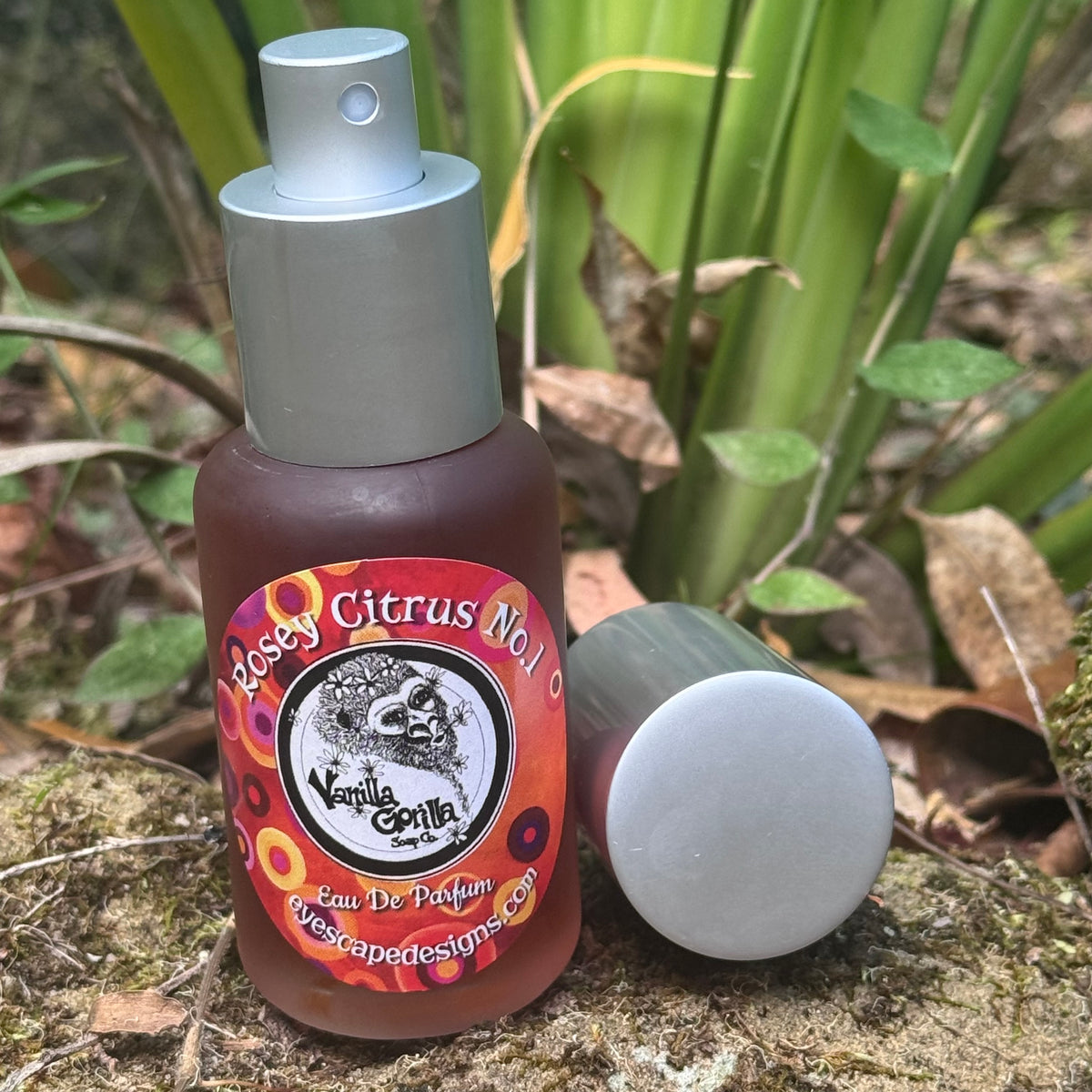A small spray bottle labeled 'Rosey Citrus No.1' by Vanilla Gorilla, shown outdoors on a rock with green leaves and dried leaves in the background. The silver spray is upright, and a matching silver cap lies beside the bottle.