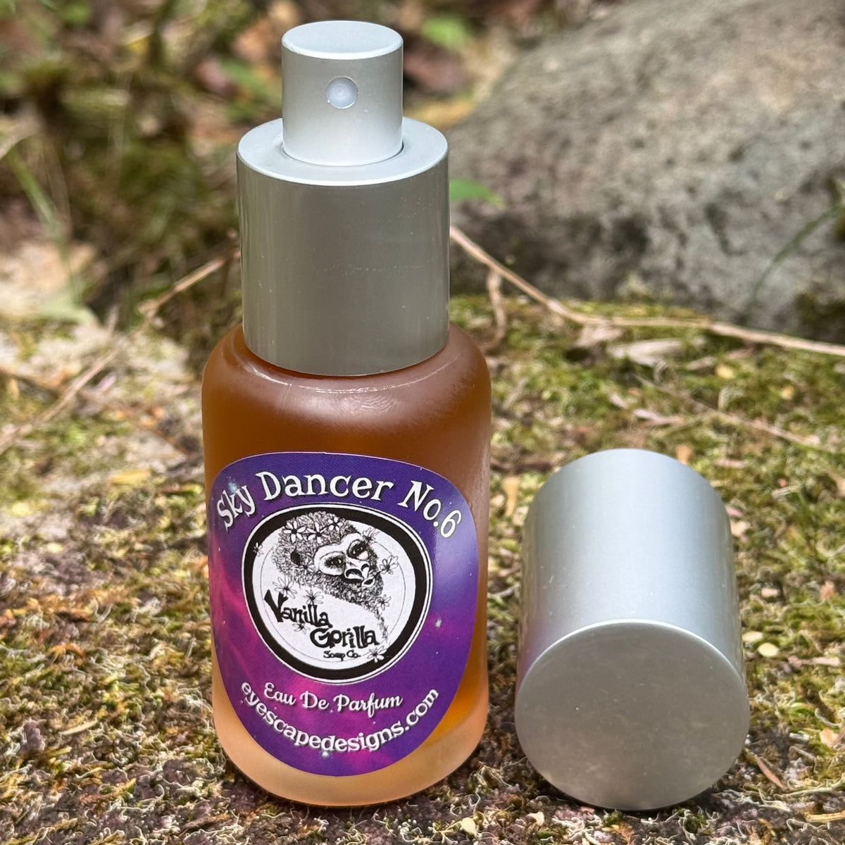 A bottle of 'Sky Dancer No. 6' Vanilla Gorilla Eau De Parfum with a purple label and silver pump, shown outdoors next to its detached silver cap on a mossy ground.