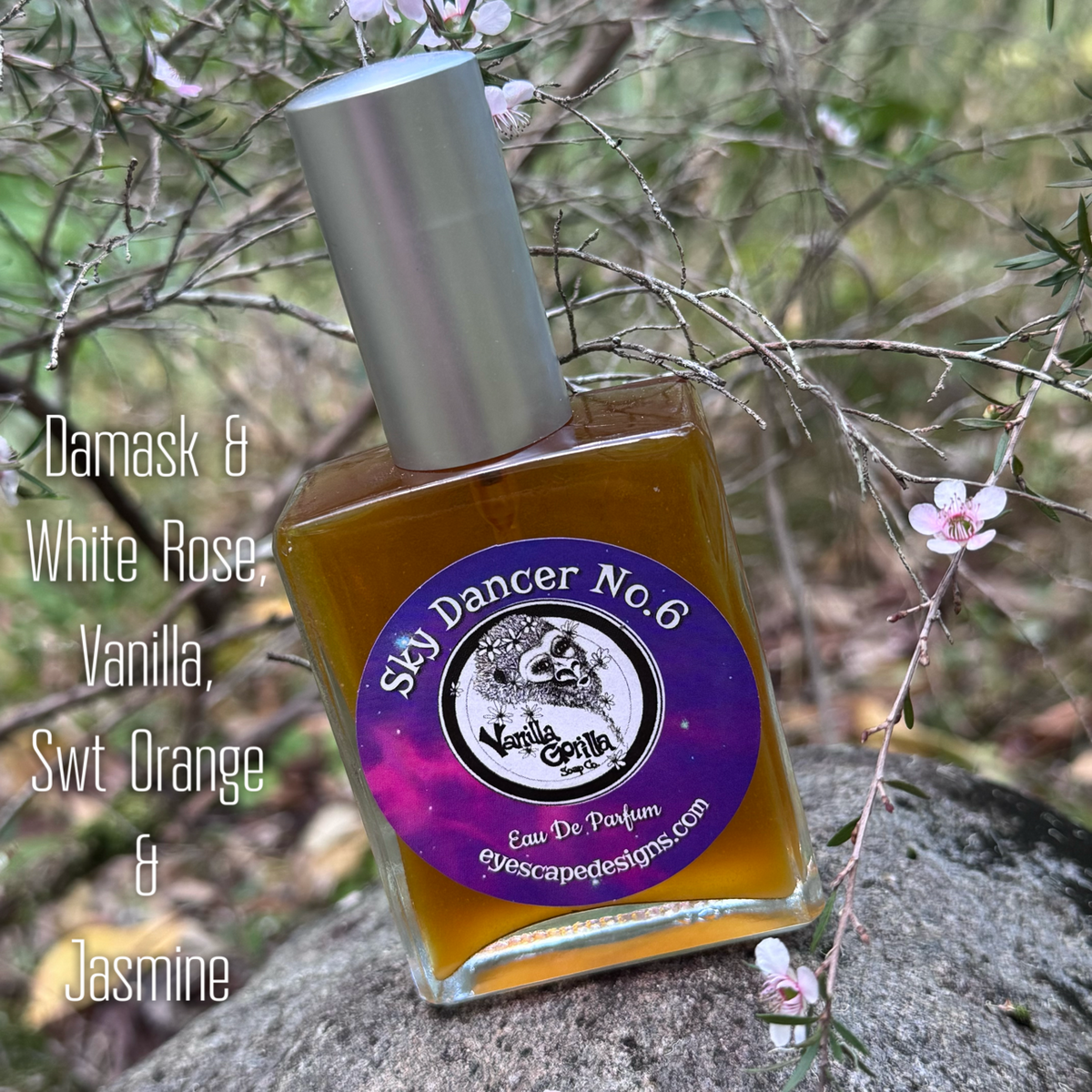 A square glass bottle of Sky Dancer No.6 Eau De Parfum with a silver cap and purple label, placed on a rock outdoors among flowering branches.