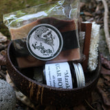 Set of natural products including a soap bar, scrubber, and jar on a stone surface with leaves.