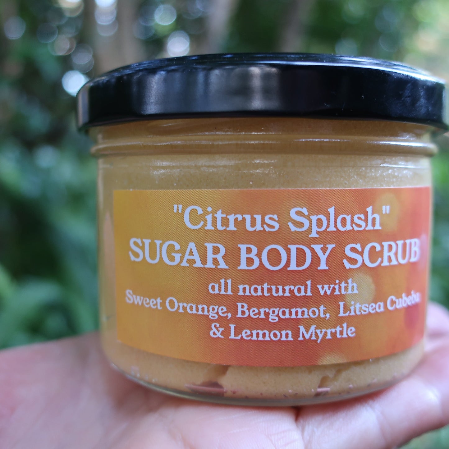Jar of 'Citrus Splash' sugar body scrub held in a hand with a blurred green background