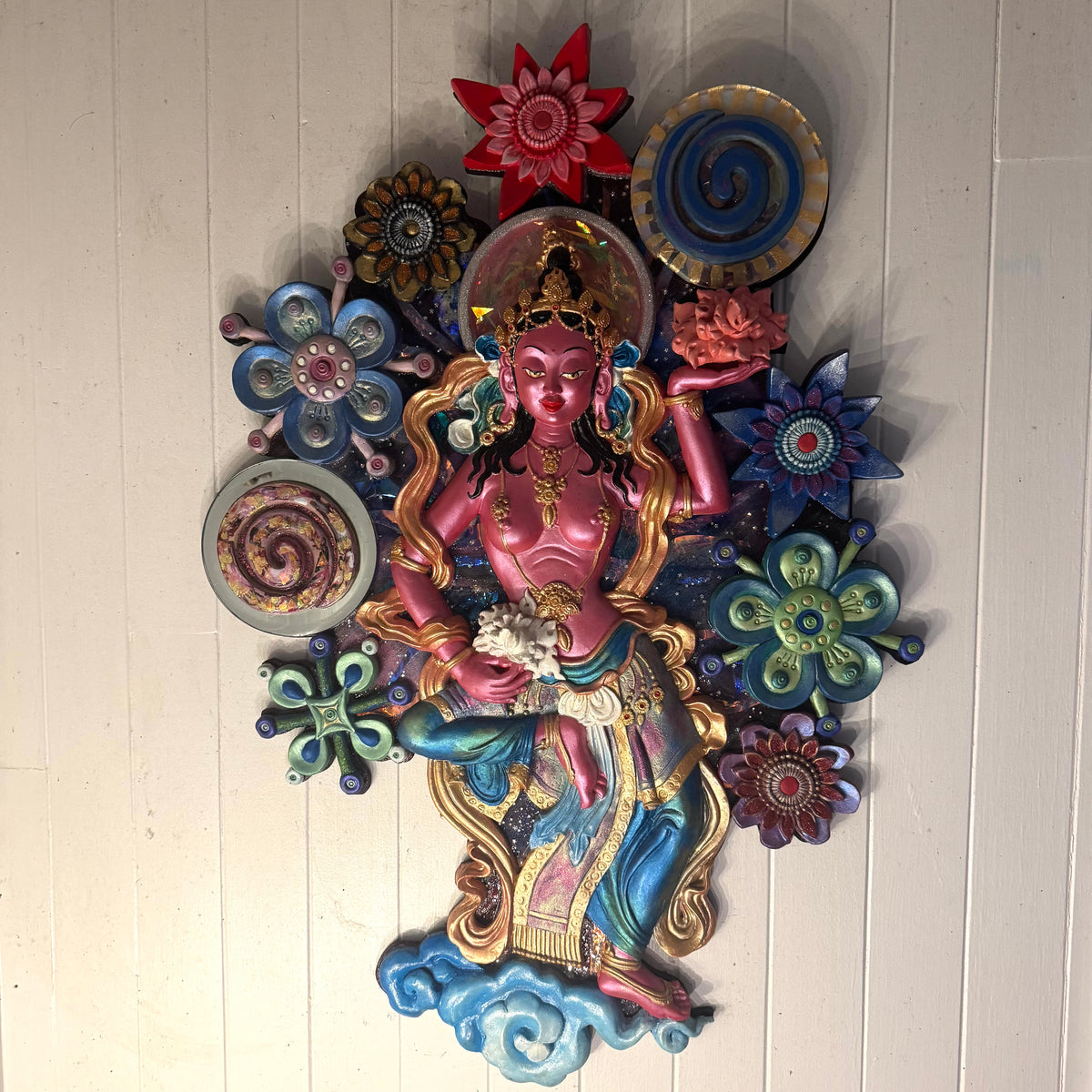 Decorative wall piece with a central figure surrounded by colorful flowers on a white wooden panel background.